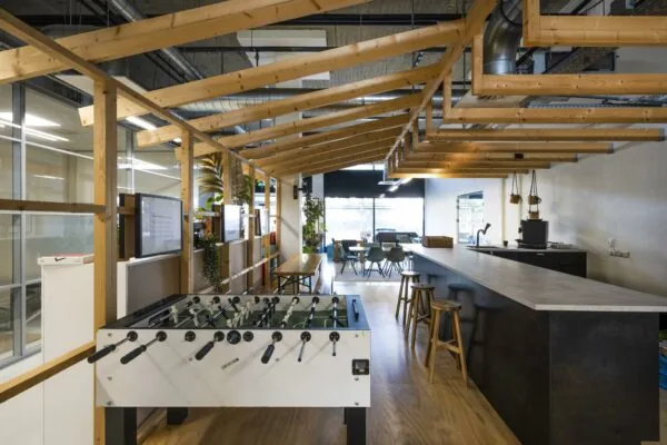 Moderne kantoorlounge aan de Joan Muyskenweg met een tafelvoetbaltafel, bar en zithoek onder een houten dakconstructie.